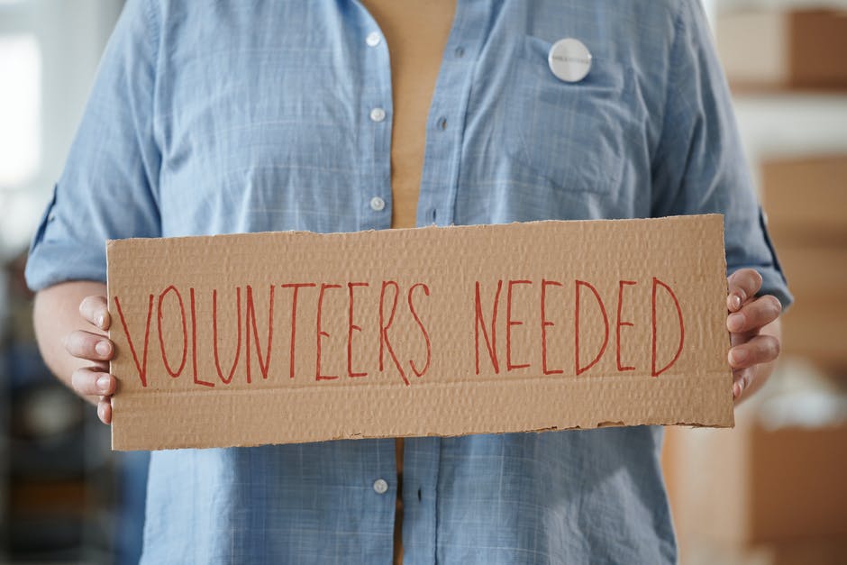 A person in a light blue long sleeve shirt holds a cardboard sign with [TEXT]: volunteers needed, written in red ink.